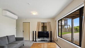 Ein Wohnzimmer mit grauem Sofa, einem Holztisch, einem Flachbildfernseher auf einem Standfuß und einem großen Fenster mit Vorhängen.