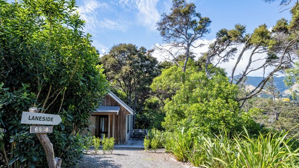 Une cabane en bois, accessible par un chemin de gravier, entourée d’une végétation luxuriante et d’arbres.