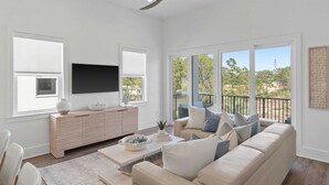 A spacious living room with a large window, a ceiling fan, a flat-screen TV, a beige sofa, and a wooden console table.