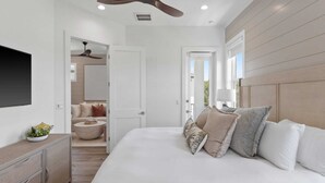 A bedroom with a large bed, a ceiling fan, a TV mounted on the wall, and a view of the outdoors through a door.
