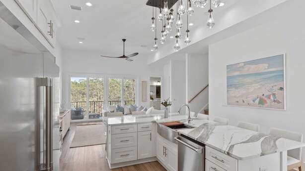 A modern kitchen with white cabinetry, a marble countertop, and a large island. There is a ceiling fan, pendant lights, and a painting of a beach scene on the wall.