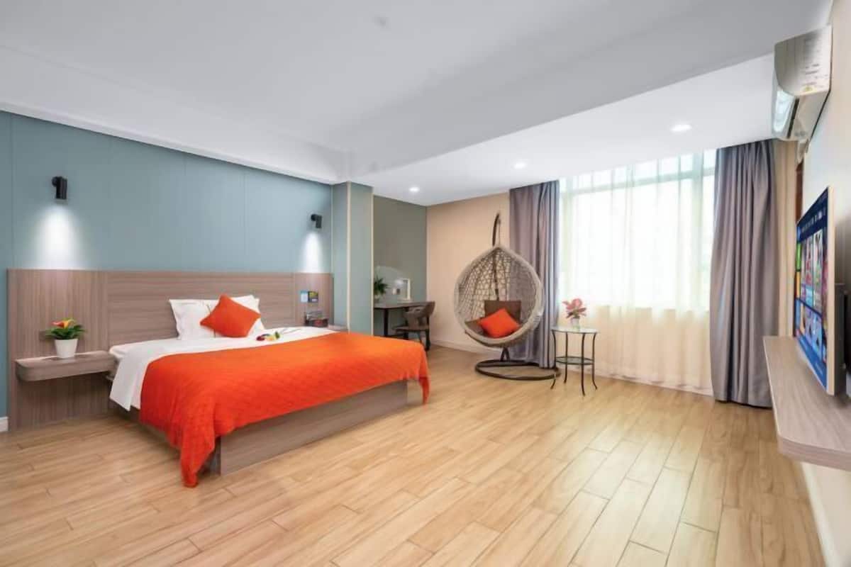 Una habitación de hotel moderna con suelo de madera, una cama con colcha naranja, un sillón colgante, una mesita con lámpara y un ventanal con cortinas.
