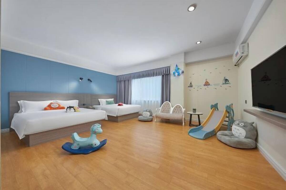 Habitación de hotel con cama, sofá, zona de juegos con juguetes, televisor y ventana con cortinas.