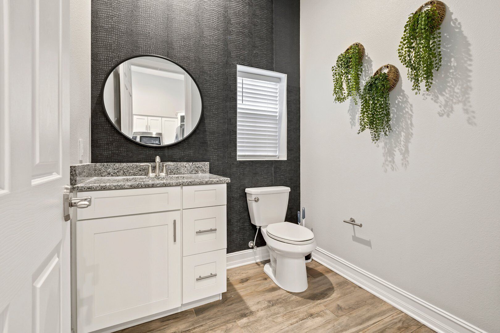 A bathroom with a white vanity, a round mirror, a toilet, and a window with blinds.