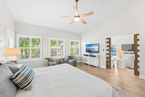 A bedroom with a bed, a ceiling fan, a TV, and a view of the outdoors through large windows.