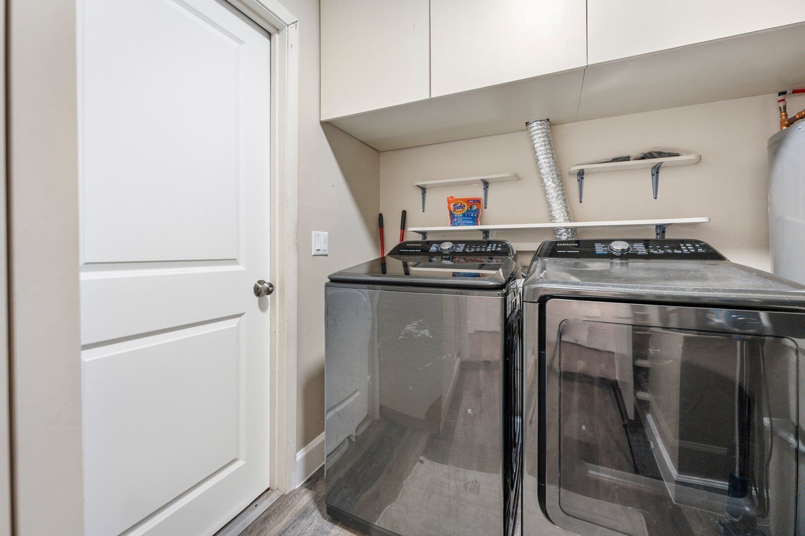 A laundry room with two front-loading washing machines, a shelf with cleaning supplies, and a vent above the machines.