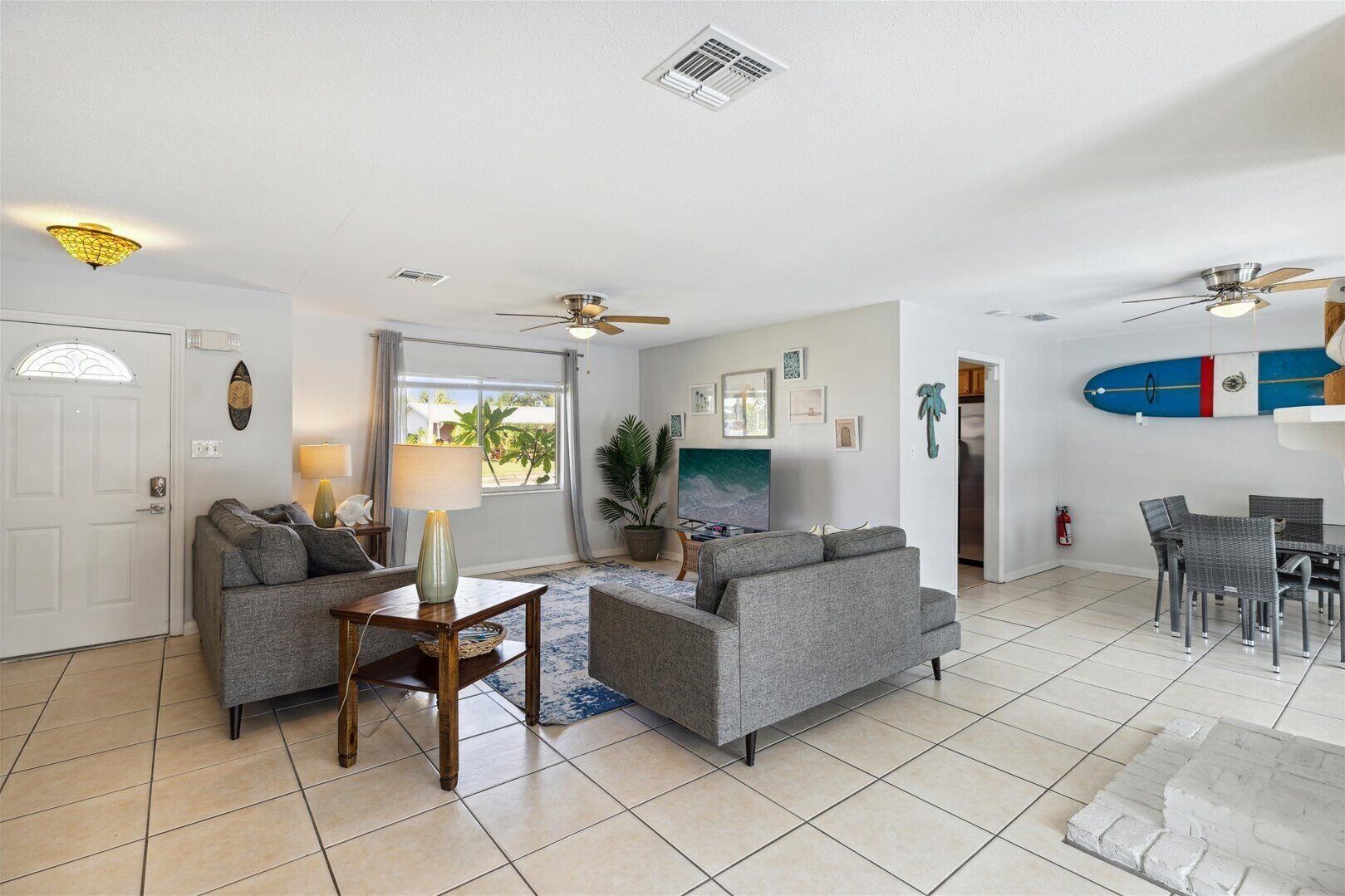 A spacious living area with two grey sofas, a wooden coffee table, a ceiling fan, a TV, and a surfboard on the wall.