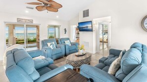 A spacious living room with a large window offering a view of the beach, a ceiling fan, and a flat-screen TV mounted on the wall. - 3435 S Highway A1A (Melbourne Beach)