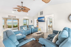 A spacious living room with a large window offering a view of the beach, a ceiling fan, and a flat-screen TV mounted on the wall.