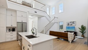 A spacious kitchen with white cabinetry, a central island, and a stainless steel refrigerator. A staircase leads to a loft area with a view of the outdoors. - 132 Ocean Estates Dr (Fort Pierce)