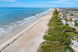 An aerial view of a beach with a long stretch of sand, adjacent to a residential area with houses and greenery. - 221 Cherie Down Ln (Cape Canaveral)