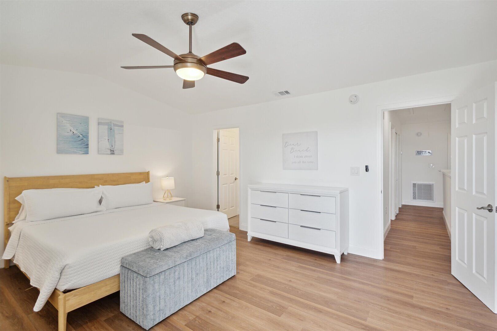 A bedroom with a wooden bed, a white dresser, a bench, a ceiling fan, and a hallway leading to a door.
