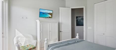 Ein Schlafzimmer mit einem Bett, einem weißen Kleiderschrank, einem weißen Sessel und einem an der Wand befestigten Fernseher.