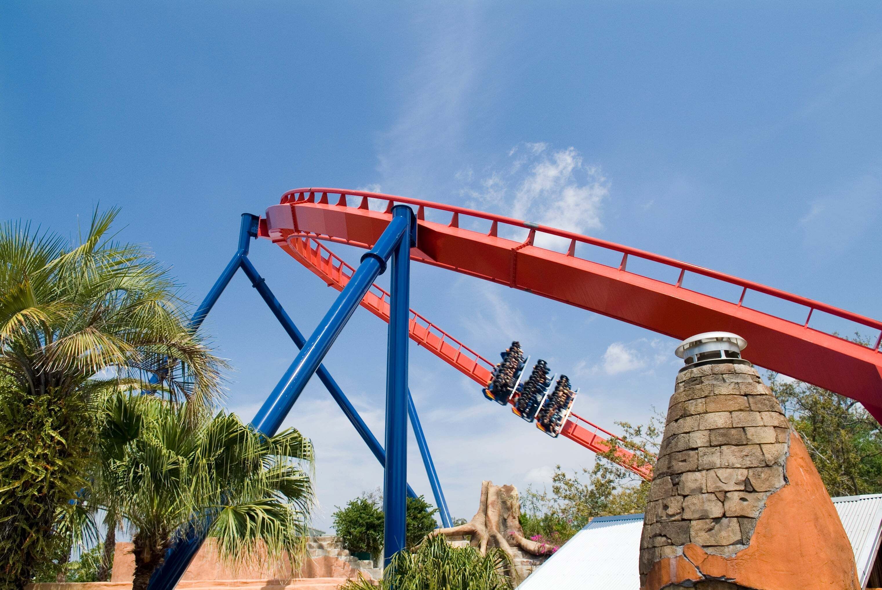 A roller coaster with a red track and blue supports, surrounded by palm trees and a stone chimney.