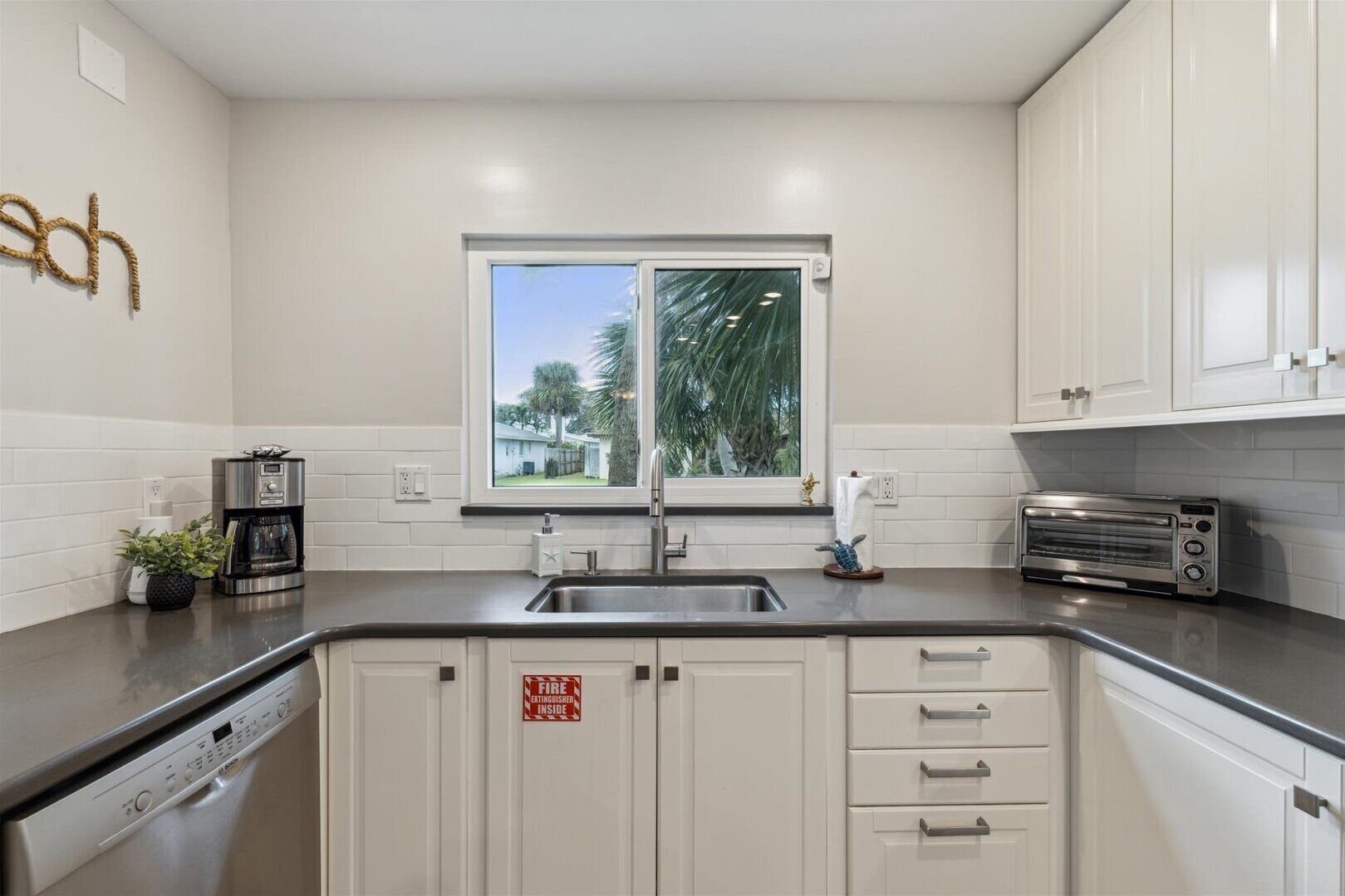 A modern kitchen with white cabinets, a stainless steel sink, a coffee maker, a toaster, and a window with a view of greenery.