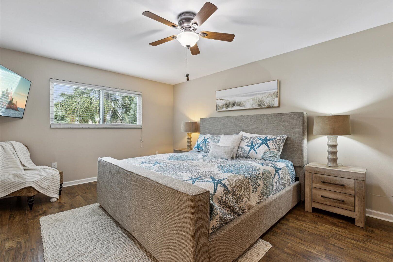 Une chambre à coucher comprenant un lit, une table de chevet, une chaise, un ventilateur de plafond et une fenêtre avec des stores.