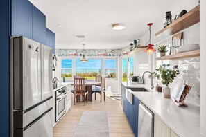 A modern kitchen with a view of the beach, stainless steel appliances, wooden chairs, and a dining table.