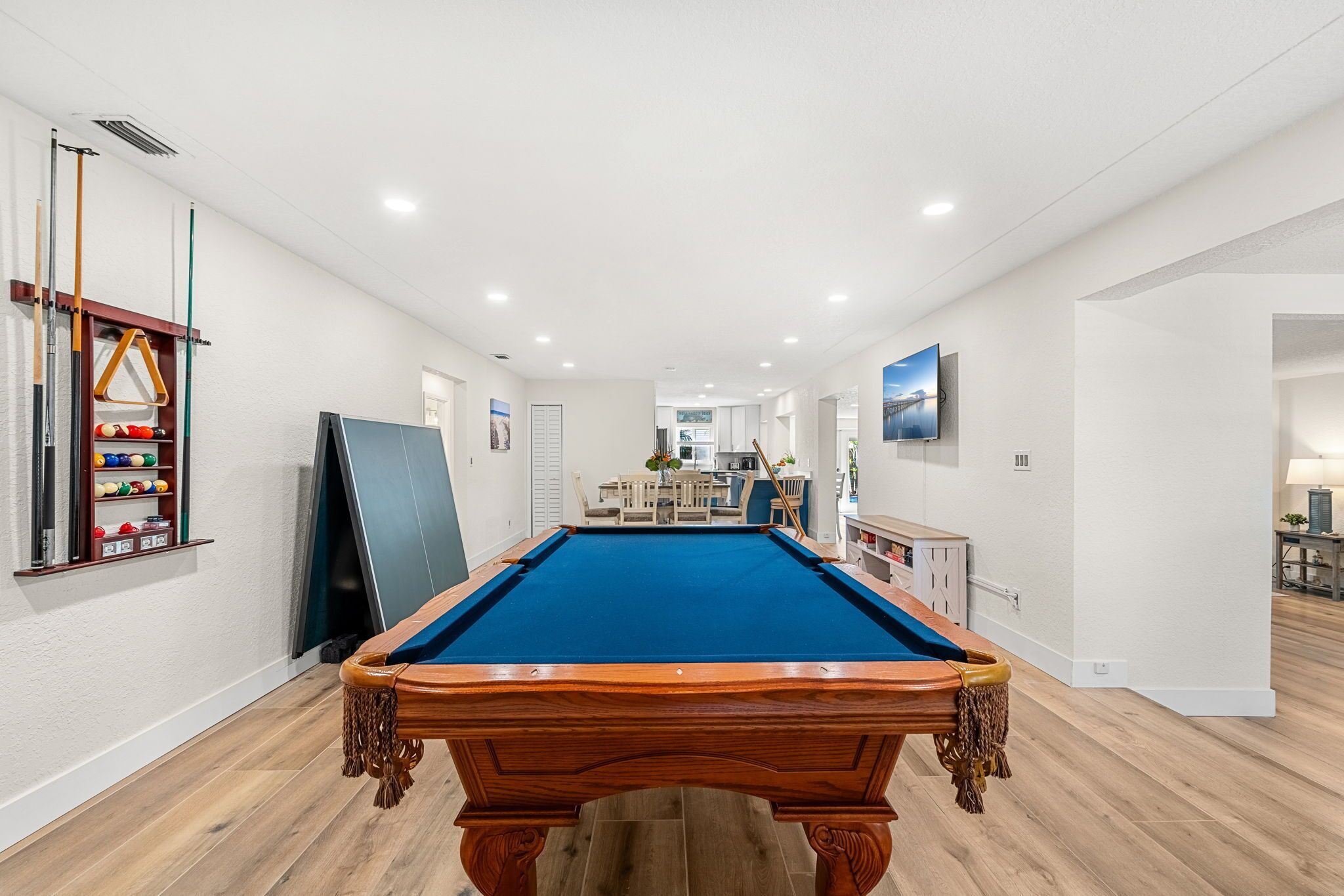 Une table de billard se trouve dans une pièce bien éclairée, dotée d’un plancher en bois, d’un porte-billes et d’un téléviseur fixé au mur.