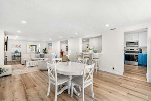 A modern living room with a white sofa, a dining table with white chairs, a kitchen with blue cabinets, and a wooden floor. - 121 Atlantic Blvd (Indian Harbour Beach)