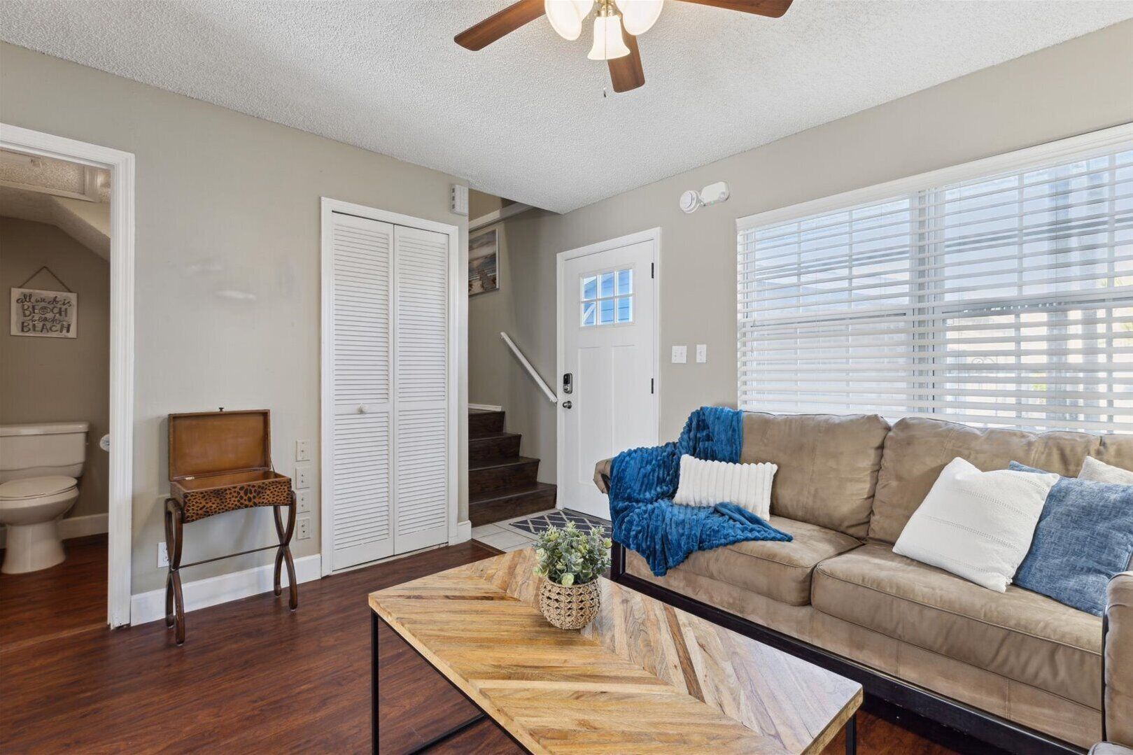 A living room with a wooden coffee table, a beige sofa with blue and white cushions, a wooden chair, and a staircase leading to another room.