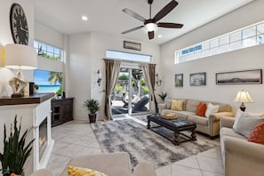 A spacious living room with a ceiling fan, a fireplace, a television, and a large window with curtains. - 106 Mediterranean Way (Indian Harbour Beach)