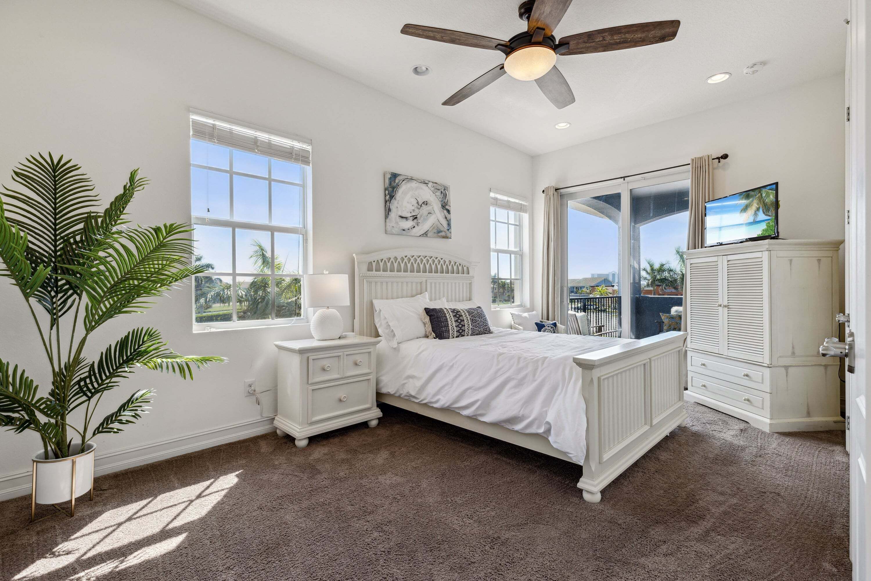 A bedroom with a bed, bedside tables, a ceiling fan, a large window, and a view of the outdoors.