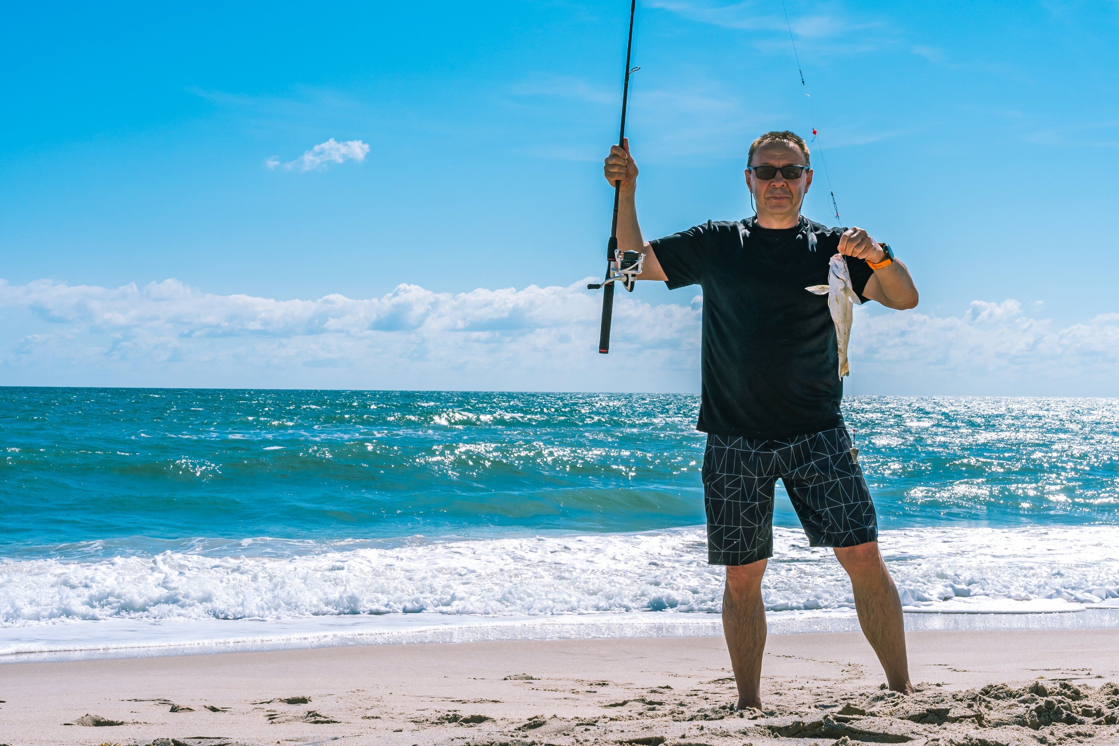 A person standing on a beach holding a fishing rod and a fish.