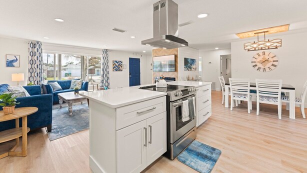 A modern kitchen with a central island, white cabinetry, and stainless steel appliances. There is a dining area with white chairs and a table, and a living room with a blue sofa and a patterned rug. - 384 Harbor Drive (Cape Canaveral)