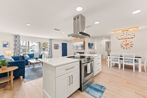 A modern kitchen with a central island, white cabinetry, and stainless steel appliances. There is a dining area with white chairs and a table, and a living room with a blue sofa and a patterned rug. - 384 Harbor Drive (Cape Canaveral)
