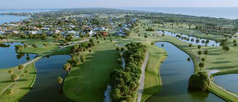 An aerial view of a golf course with water hazards, a path, and residential areas in the background.
