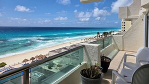 En balkon med udsigt over stranden og havet. Der er liggestole og parasoller på balkonen.