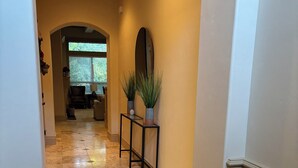 Un pasillo con suelo de baldosas, una consola con plantas en macetas, un espejo y una puerta que da a otra habitación.