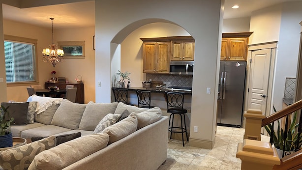 Una sala de estar con un sofá seccional color beige, un comedor con mesa y sillas, una cocina con gabinetes de madera y electrodomésticos de acero inoxidable, y una escalera con barandilla de madera.