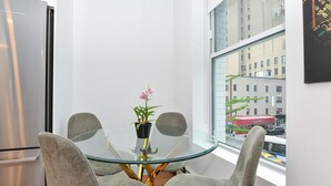 Een hoek van een moderne keuken met een glazen tafel, twee grijze stoelen, een potorkidee en een uitzicht op een stadsbeeld door een groot raam.