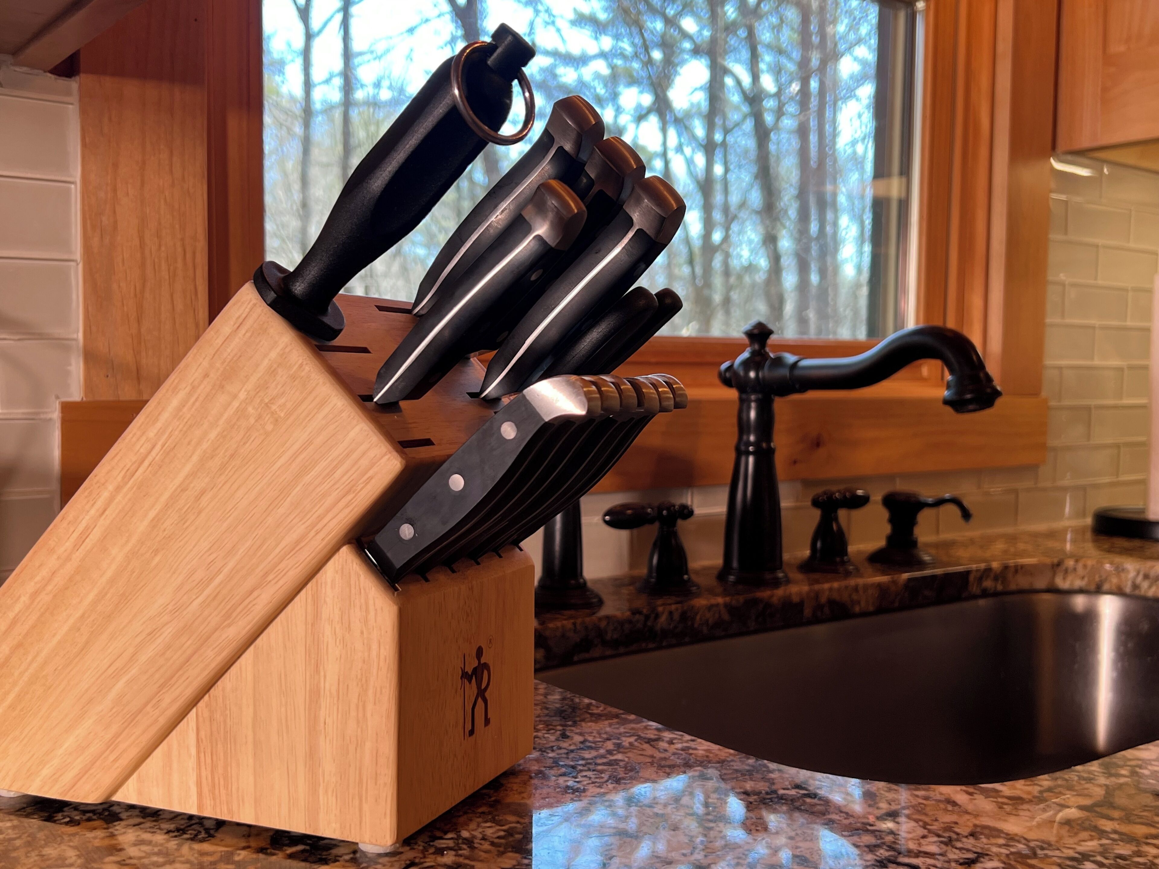 A kitchen with a wooden knife block containing several knives, a sink, and a window with a view of trees.