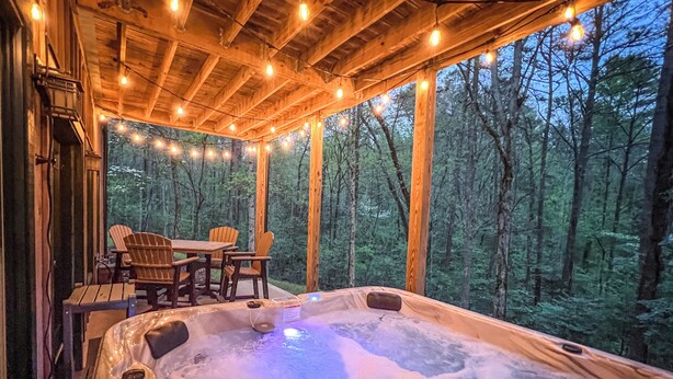 Un bain à remous avec une terrasse en bois et des guirlandes lumineuses, le tout entouré d’une forêt.