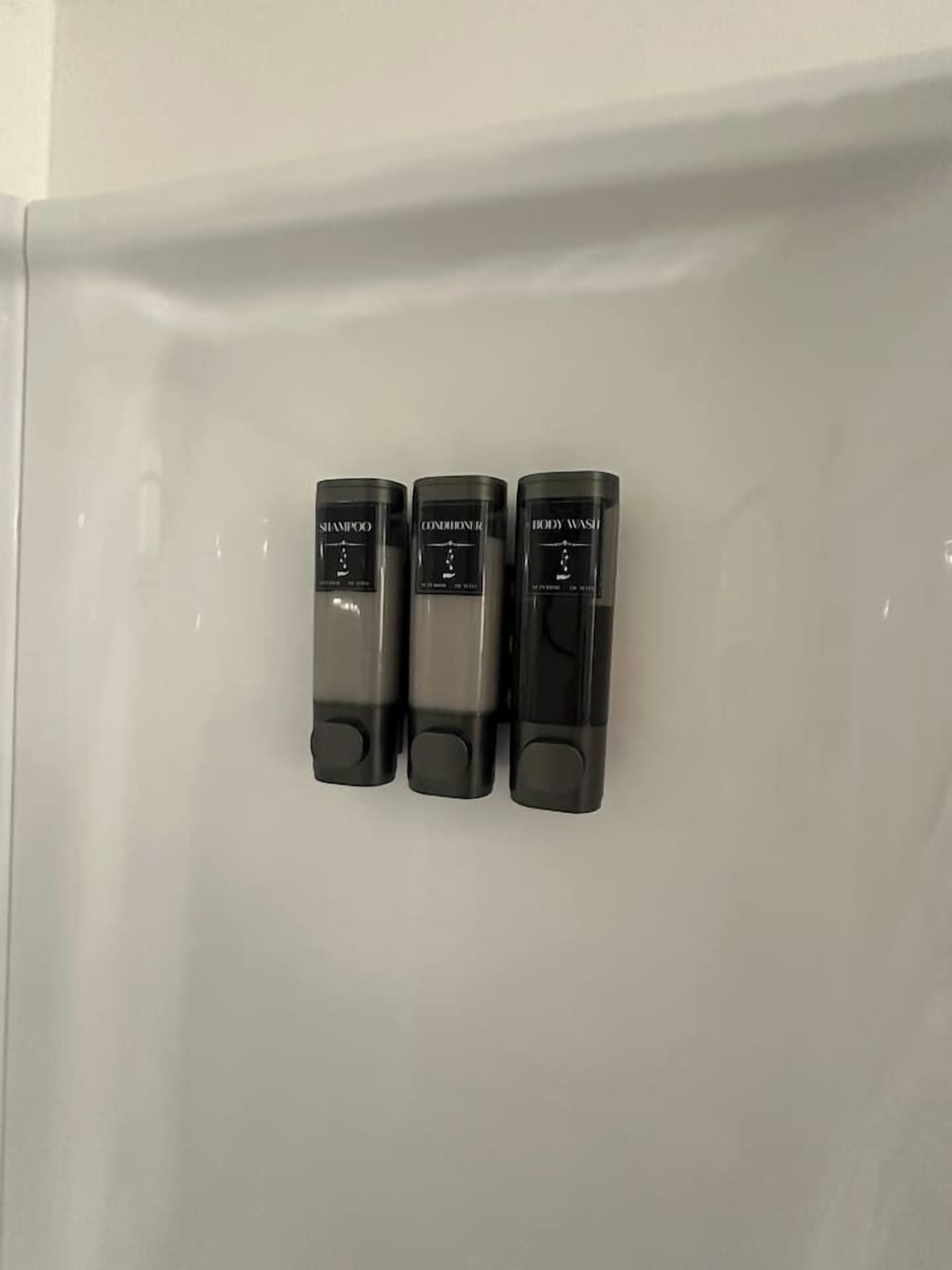 Drei schwarze Flaschen mit den Aufschriften 'Shampoo', 'Spülung' und 'Duschgel' auf einer weißen Oberfläche.