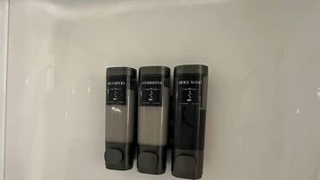 Drei schwarze Flaschen mit den Aufschriften 'Shampoo', 'Spülung' und 'Duschgel' auf einer weißen Oberfläche.