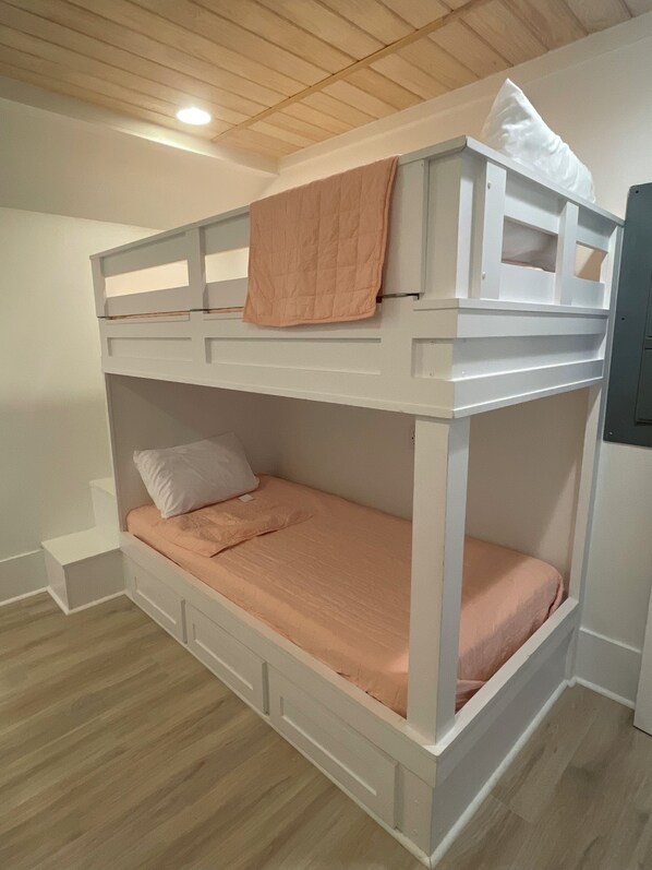 A bunk bed with a wooden headboard, a lower bed with a mattress and pillows, and a white wall with a small shelf.