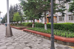 Uma passarela pavimentada com padrão xadrez, delimitada por um baixo borda vermelha e sebes verdes, conduz a um edifício com porta amarela.
