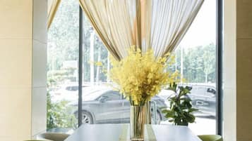 A table with a vase of yellow flowers, a window with curtains, and a view of parked cars outside.
