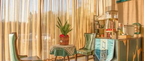 A room with a large window, two chairs, a small table with a plant, and a cabinet with various items.
