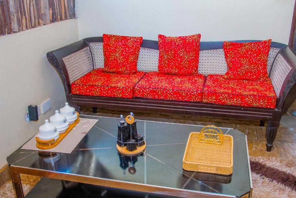Un salon avec un canapé marron foncé, des coussins à motifs rouges, une table basse en verre ornée d’un objet décoratif et un panier en osier.