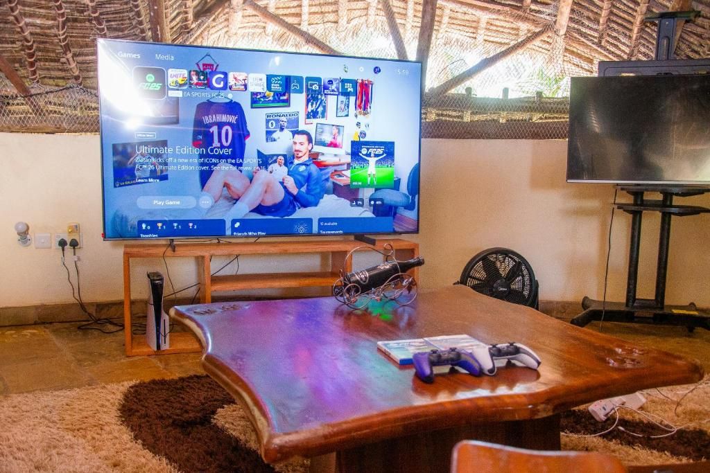 Quarto com televisão grande exibindo um jogo esportivo, mesa de madeira com microfone e um ventilador.