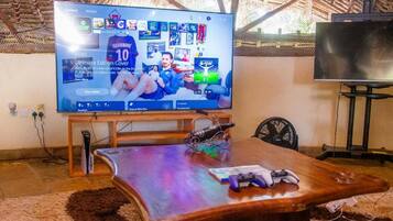 Quarto com televisão grande exibindo um jogo esportivo, mesa de madeira com microfone e um ventilador.