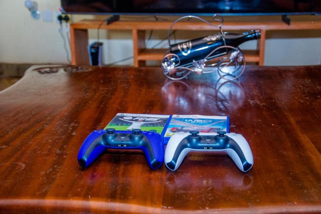 Two video game controllers on a wooden table with a game box and a guitar in the background.