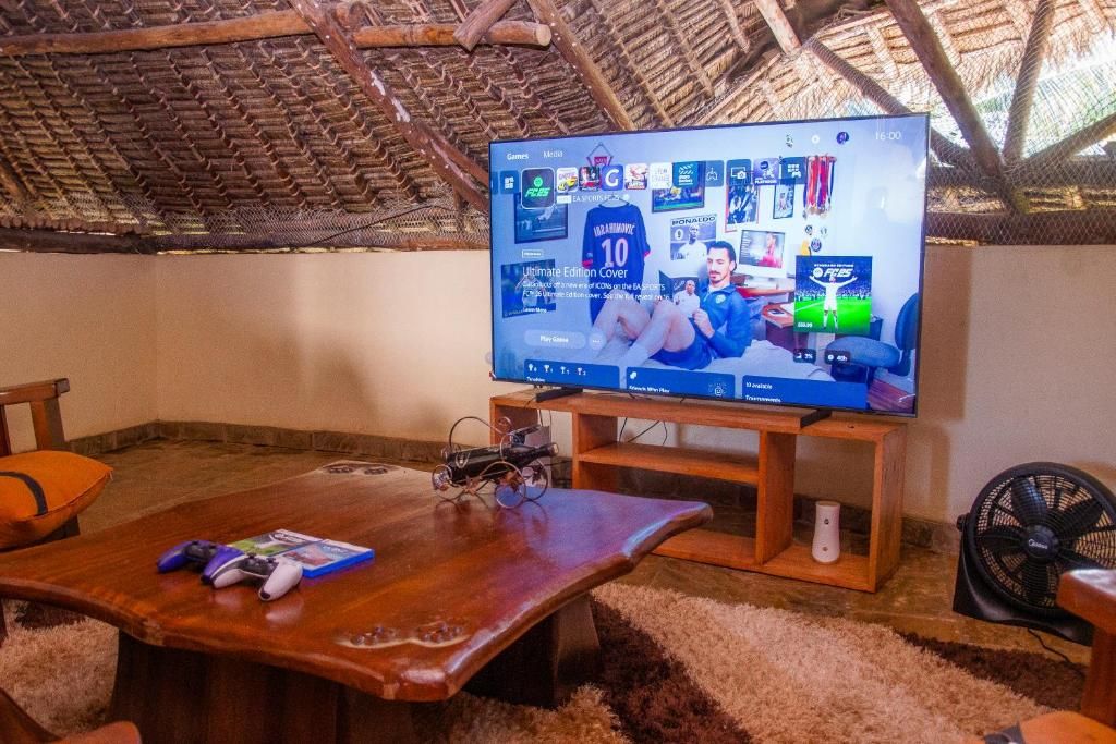 Quarto com uma grande televisão de tela plana em um suporte de madeira, uma mesa de madeira com controles de videogame e um livro, um pequeno ventilador e teto com telhado de palha.