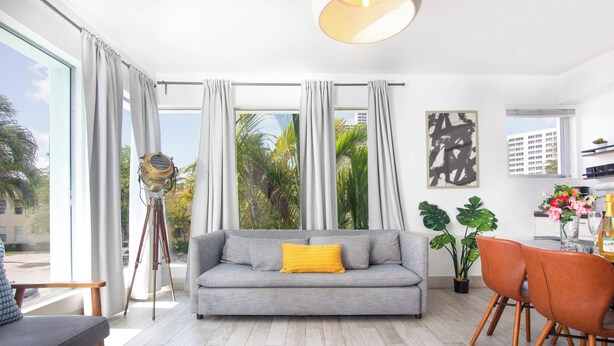 A modern living room with a grey sofa, a yellow pillow, and a dining area with orange chairs. - Boutique Apartments SoBe (Miami Beach)