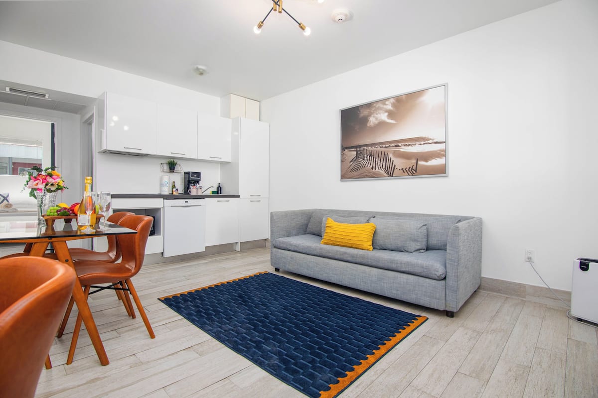 Ein modernes Wohnzimmer mit einem grauen Sofa, orangefarbenen Sesseln, einem blauen Teppich und einer Küche im Hintergrund.