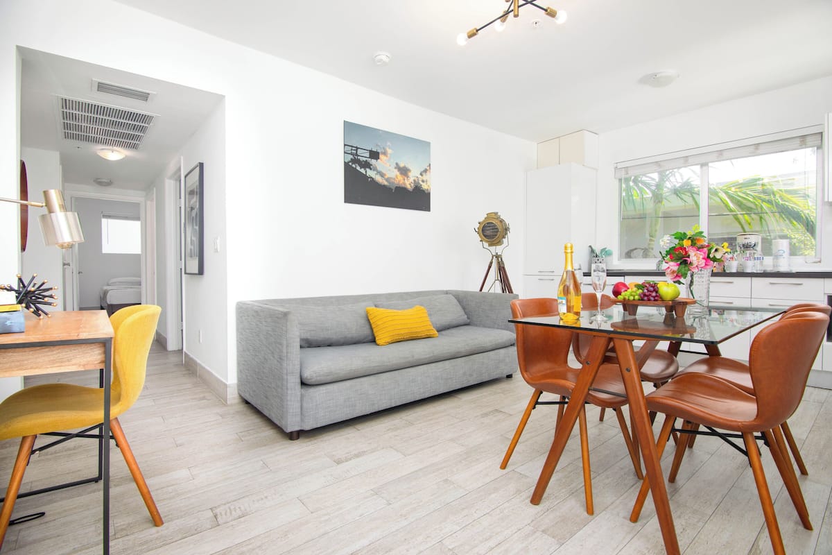 Ein modernes Wohnzimmer mit grauem Sofa, gelben und braunen Sesseln, einem Glastisch und einer Küchenzeile mit Fenster.
